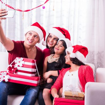 Indian Asian young family celebrating Christmas with gift while wearing Santa Hat and red Cloths sitting on white couch or sofa. Drinking, taking selfie or decorating Xmas tree. indoor